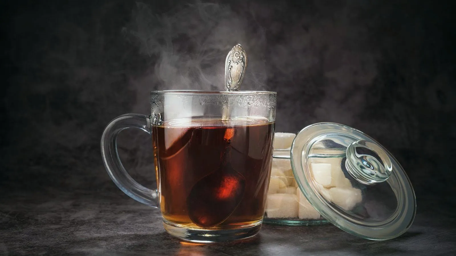 A steaming glass mug of tea with a spoon in it, beside a glass jar of white sugar cubes with the lid leaning against it.