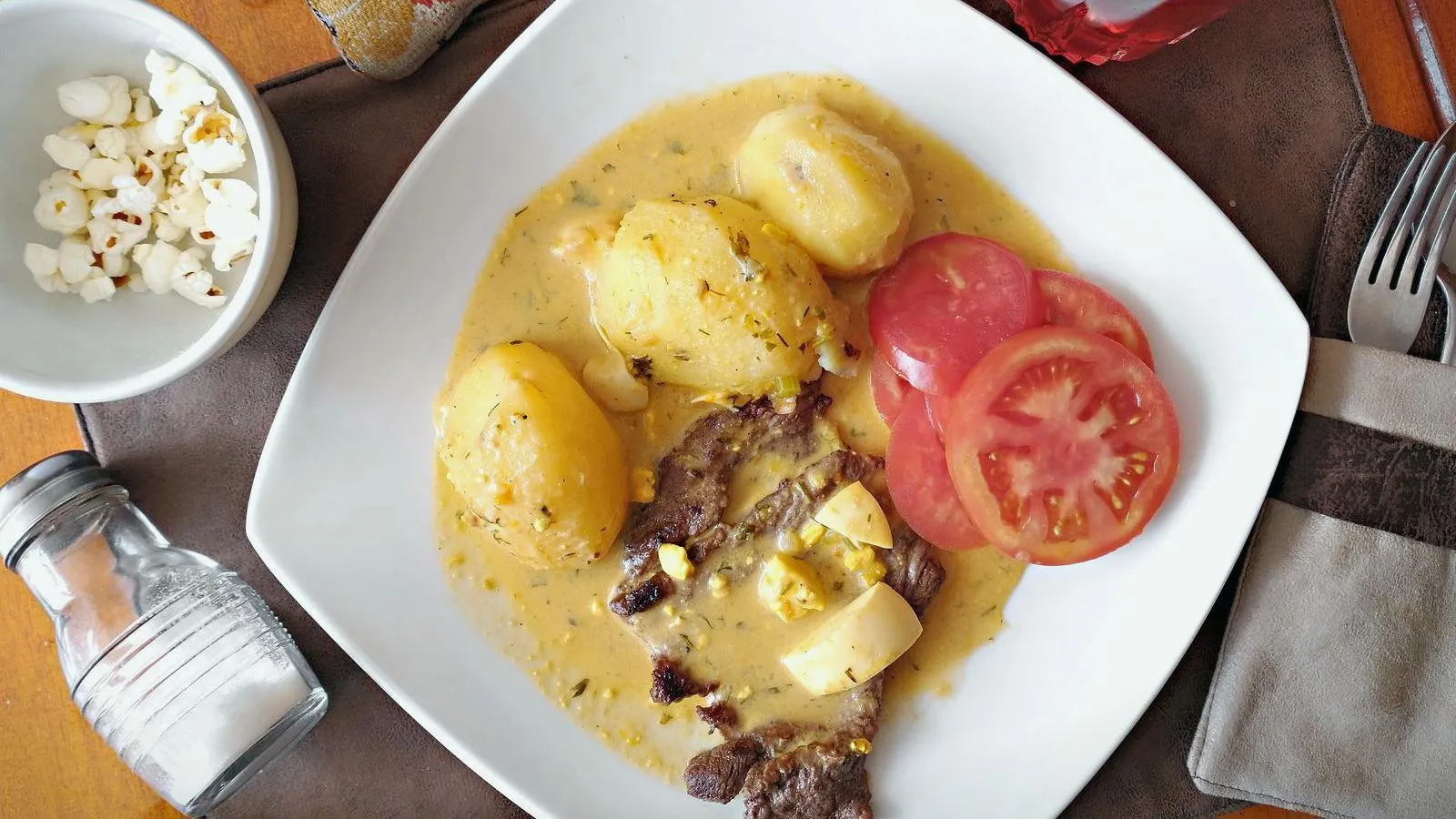 Overhead view of a plate of potatoes, sliced steak and tomato in a creamy sauce, with a glass salt shaker beside it.