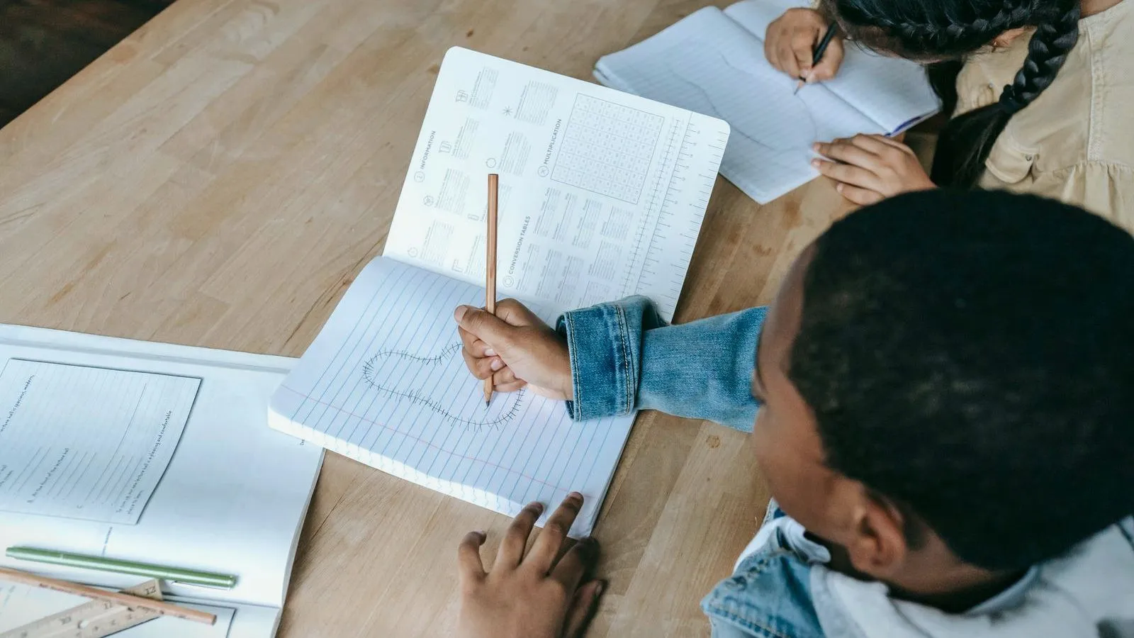 Two children write in exercise books at a wooden table, seen from above with faces out of view.