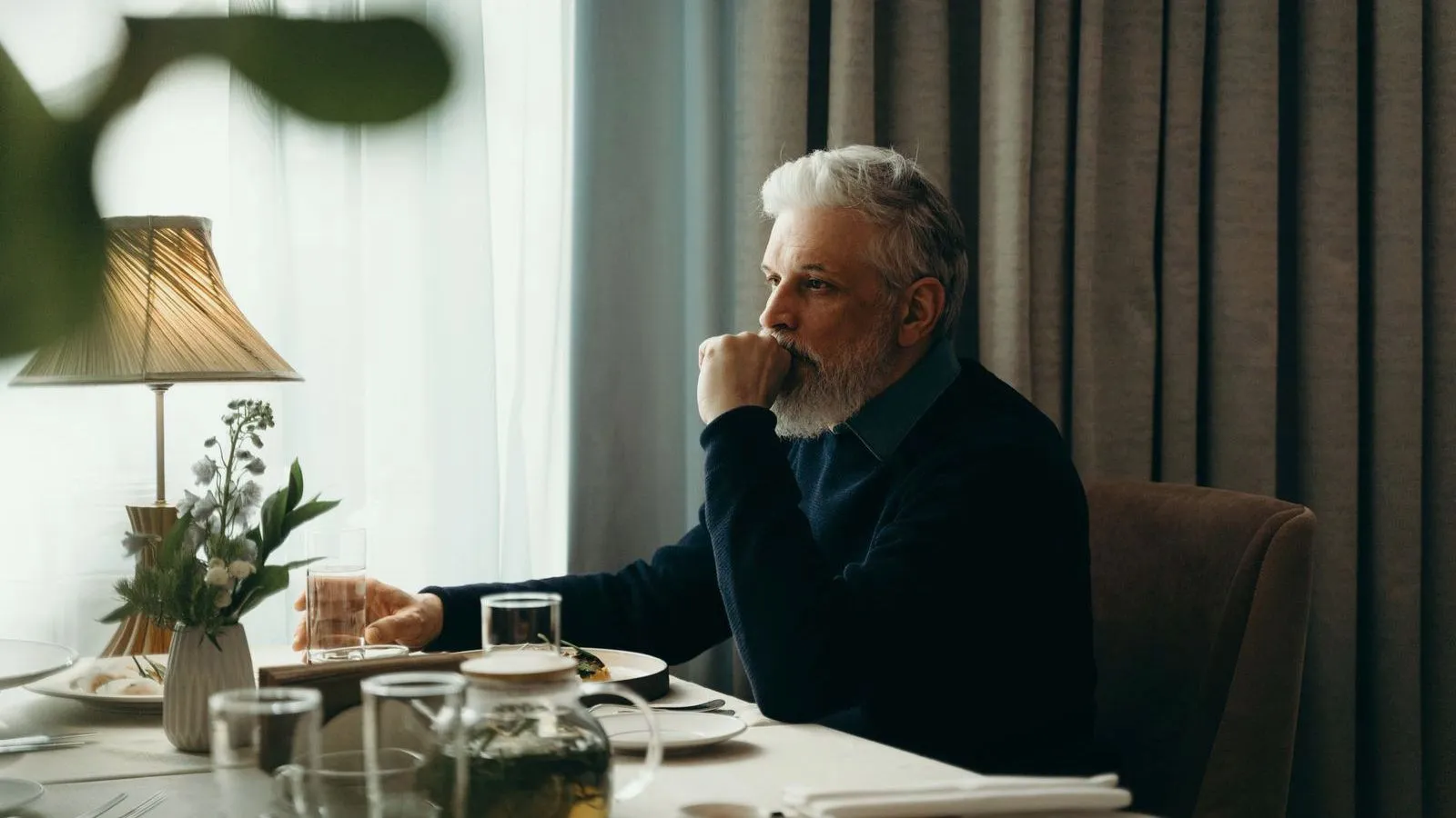 An older man with white hair and a beard sits at a laid table with a hand to the mouth, looking toward a window.