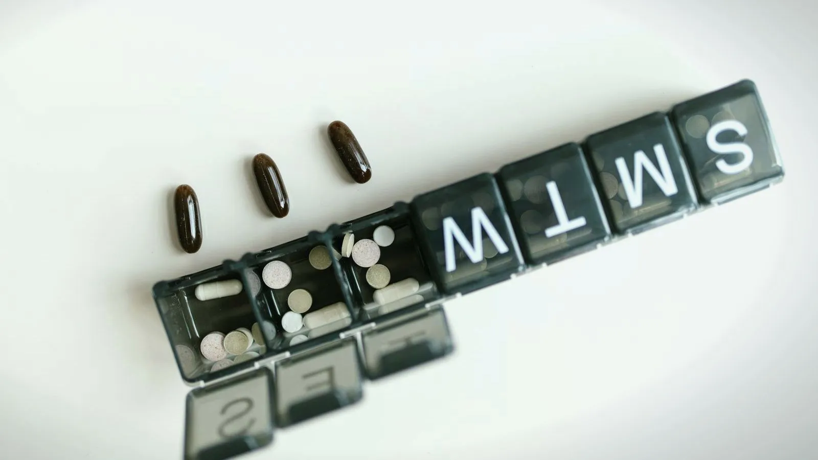 A seven-day pill organizer with three compartments open and loose tablets beside it
