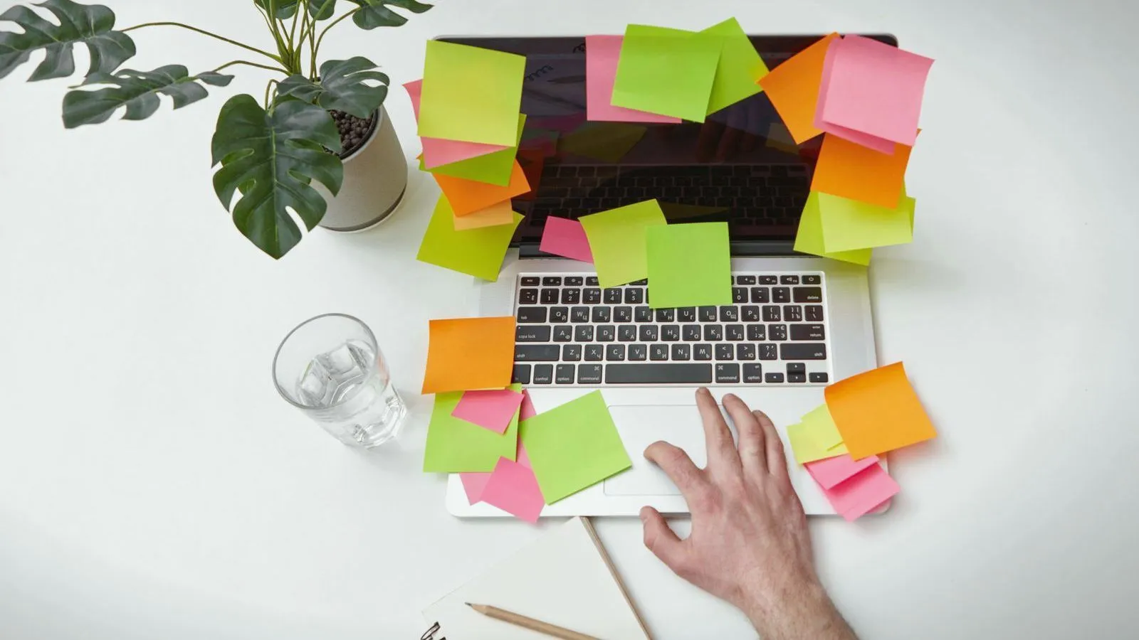 Overhead view of a laptop covered in colorful sticky notes, a hand on the keyboard, a glass of water and a potted plant on a white desk.