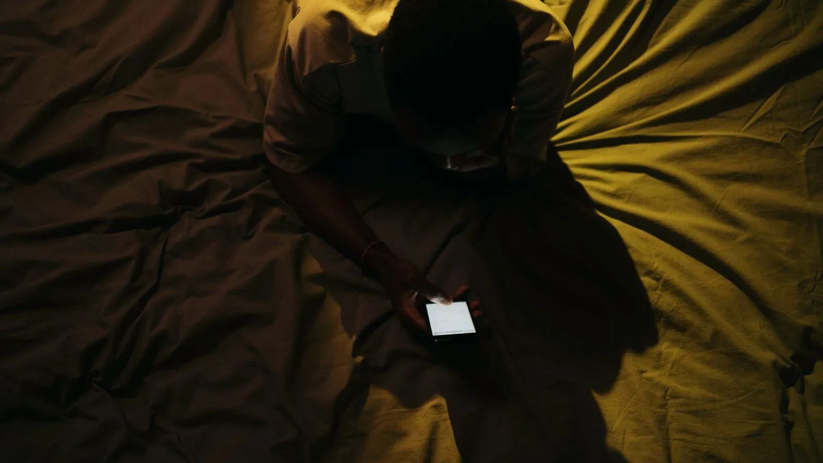 A person sits on a bed in near darkness looking down at a lit phone screen, seen from above.