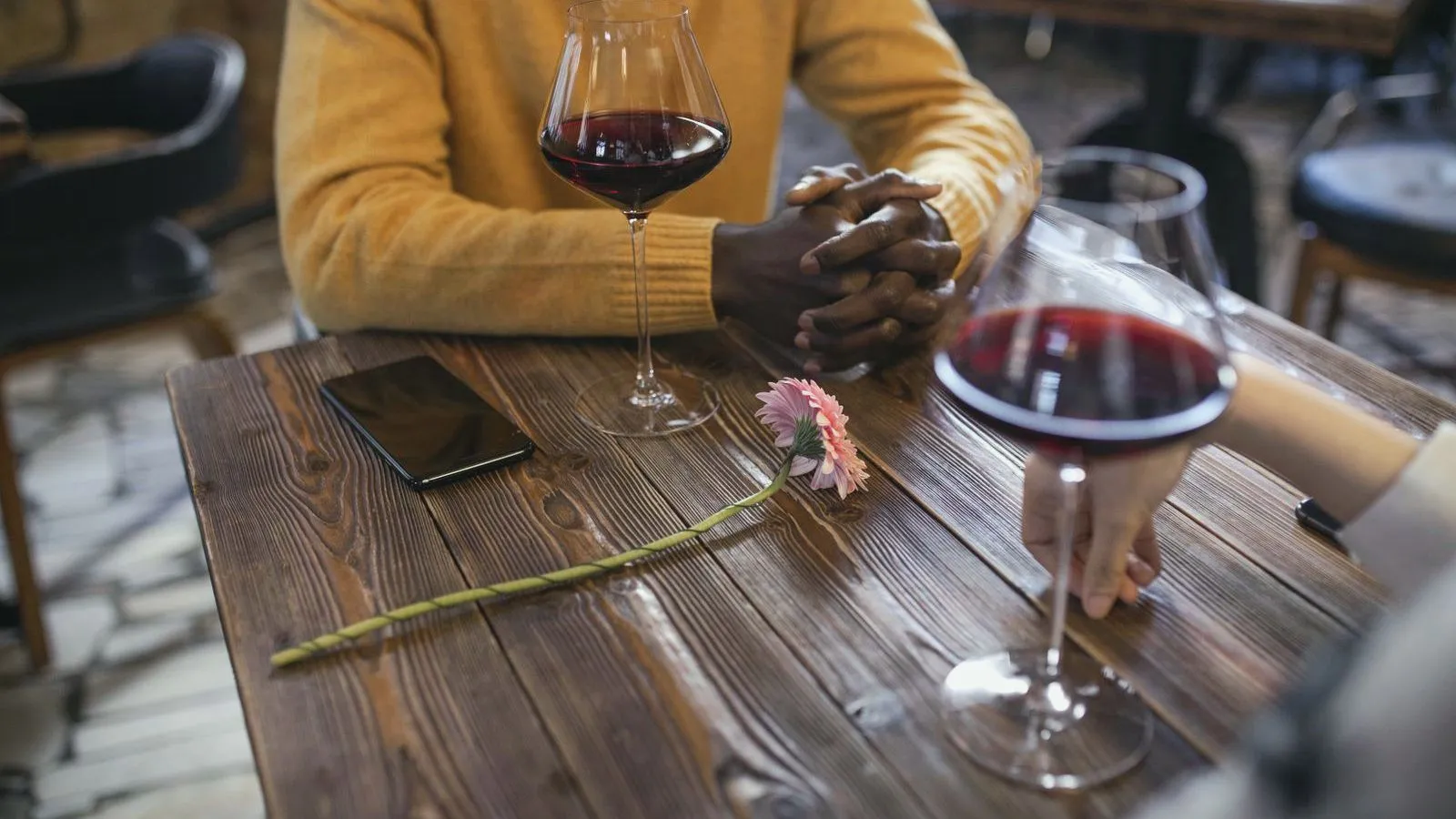 Two people's hands resting on a wooden table beside two glasses of red wine