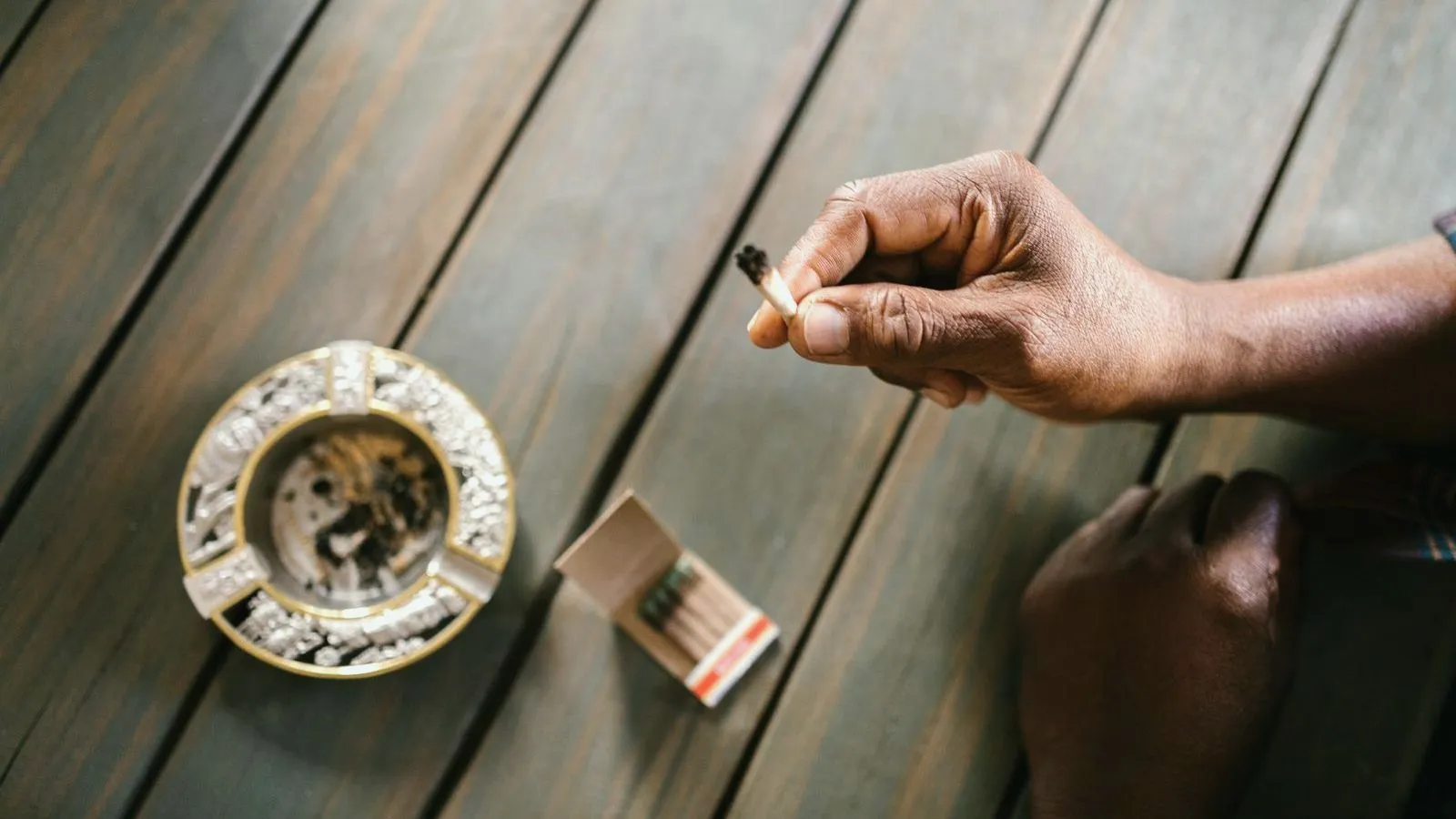 A hand holds a hand-rolled joint above a wooden table, beside an ashtray and a box of matches.