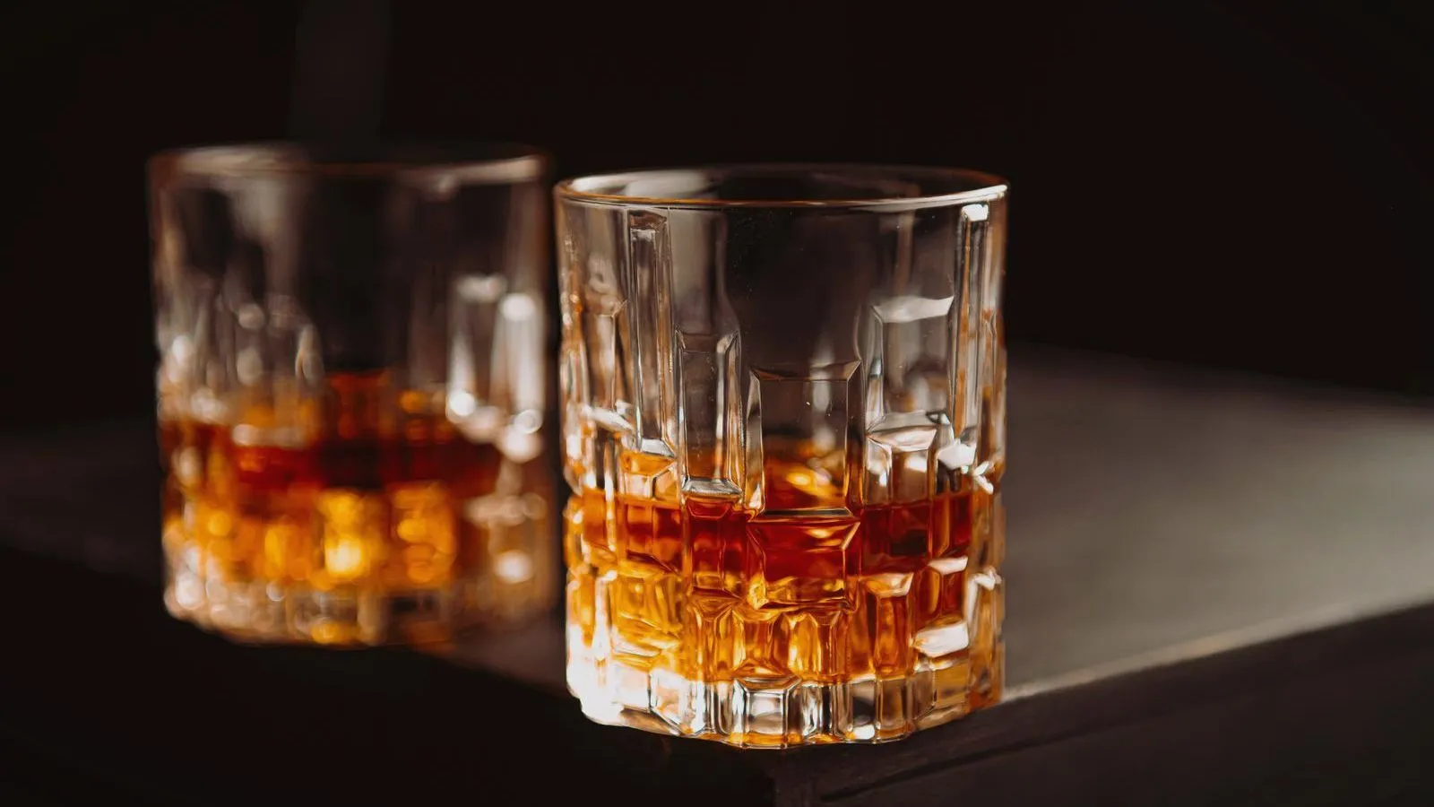 Two cut-glass tumblers of amber spirit on a dark surface.