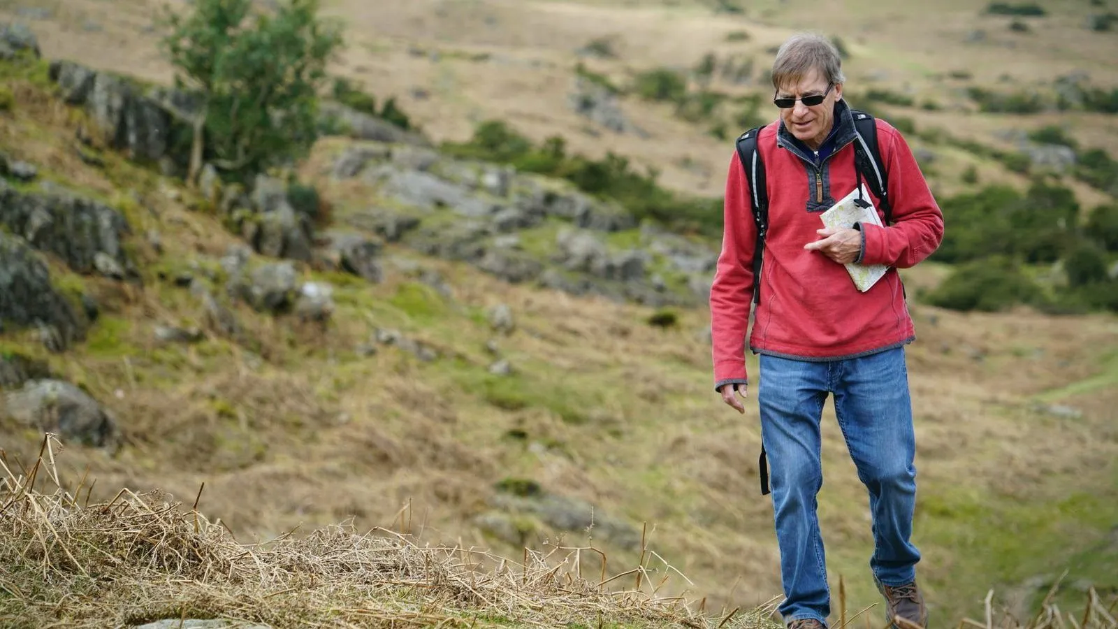 A man in a red jacket with a backpack walking across a grassy hillside