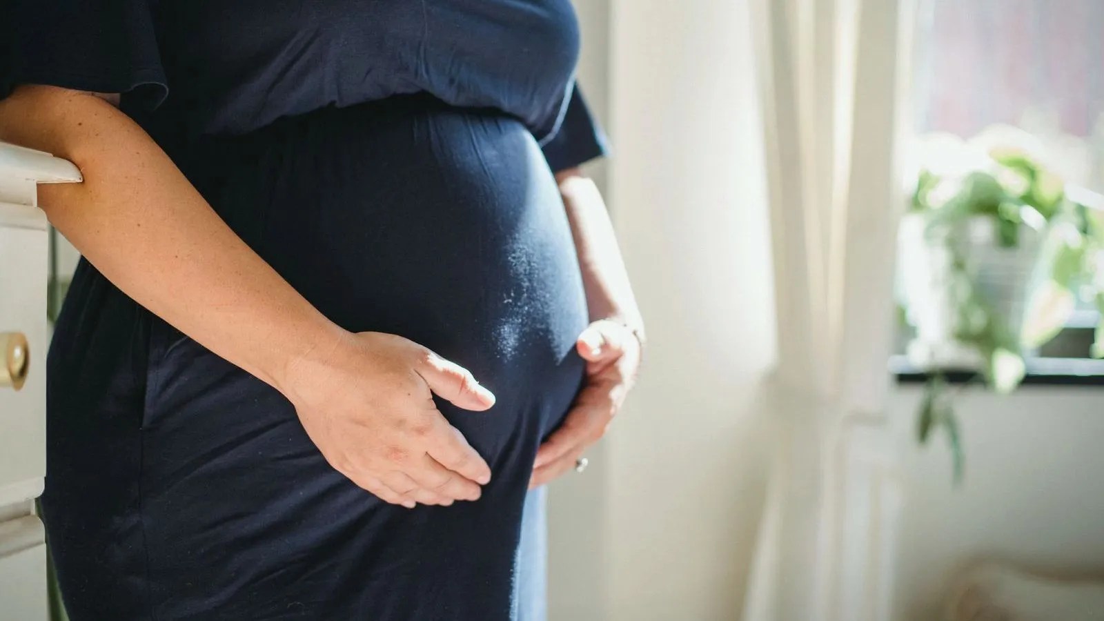A pregnant person in a navy dress stands beside a sunlit window with both hands resting on the belly, face out of frame.