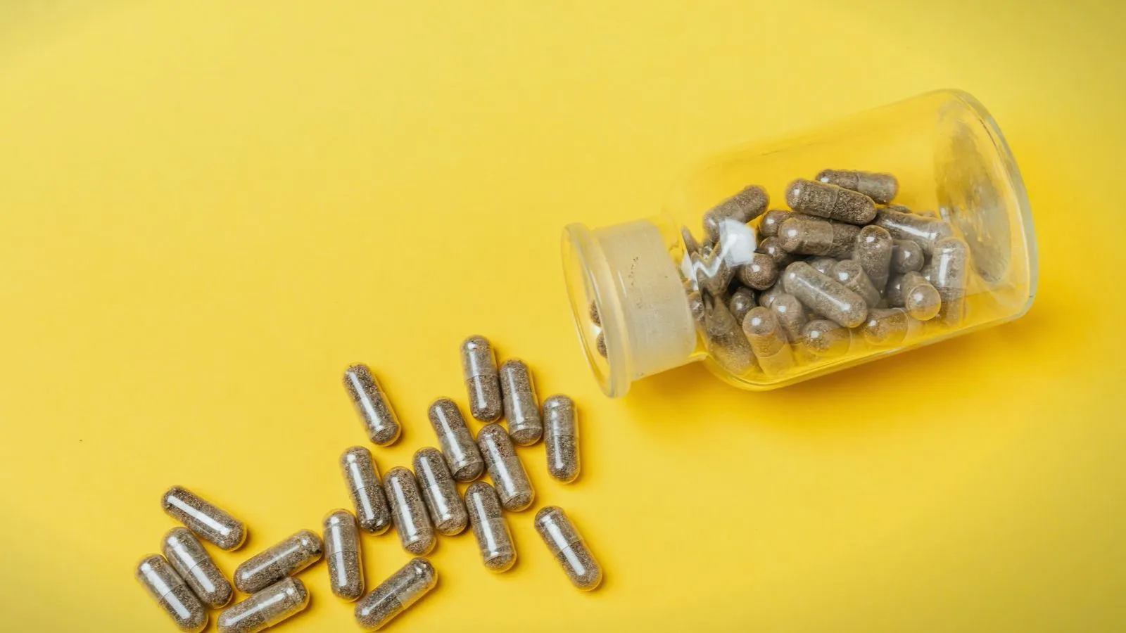 Herbal supplement capsules spilling from a glass bottle onto a yellow background