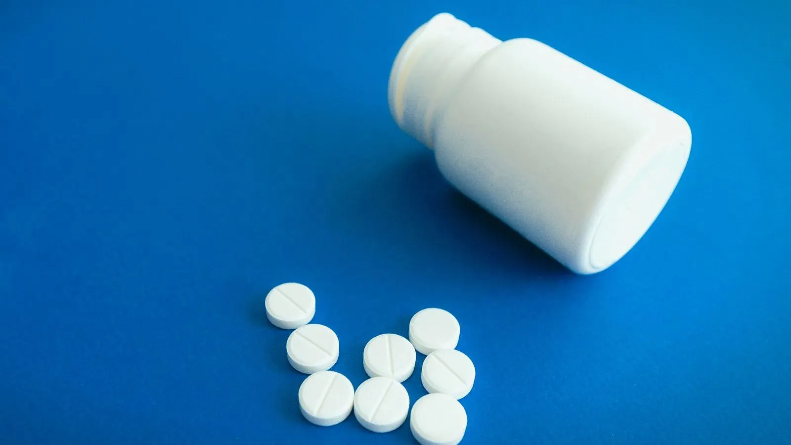 A white tablet bottle on its side with round white pills spilling onto blue