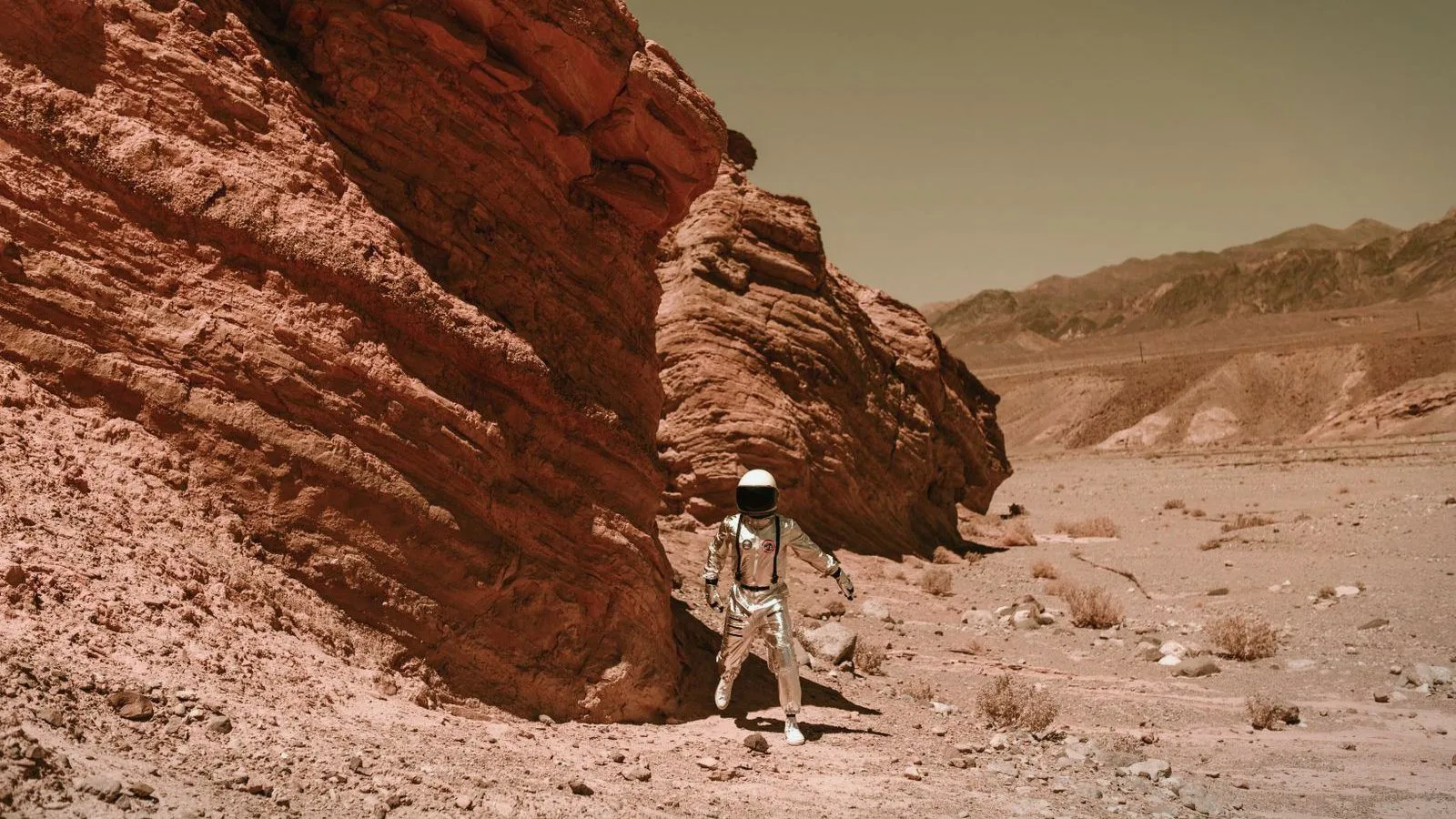 A person in a silver spacesuit walking past red rock in a barren desert