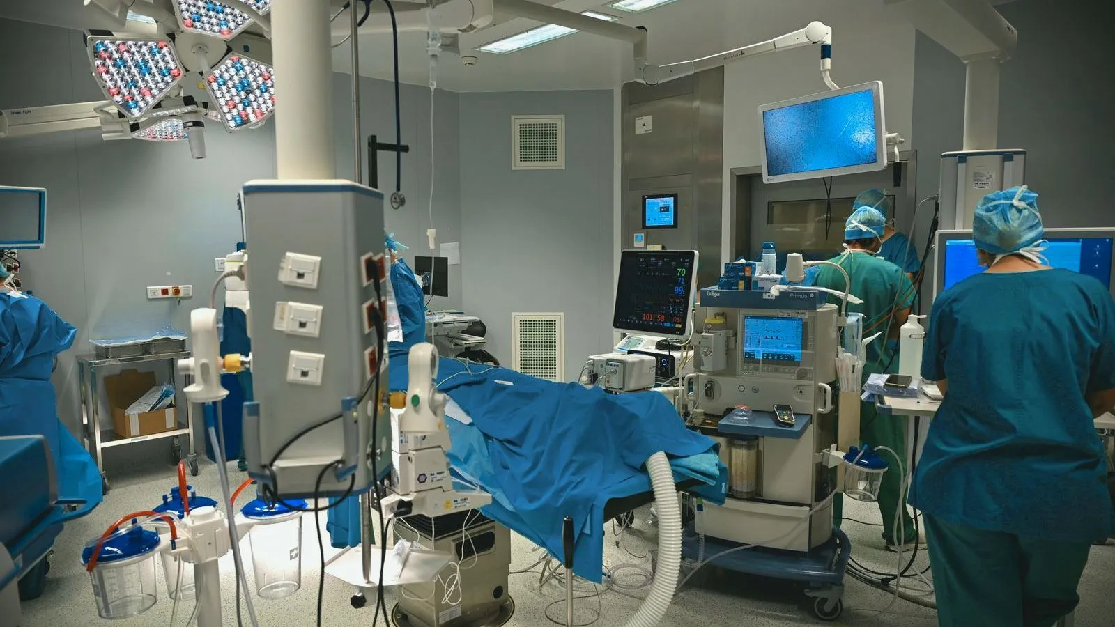 A hospital procedure room with monitors and equipment around a draped table, and staff in blue scrubs and caps seen from behind.