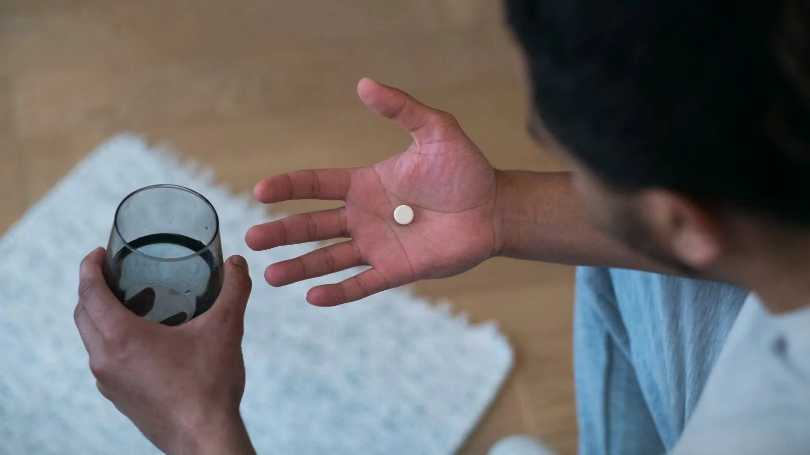 An open palm holding a single white tablet, with a glass of water in the other hand, seen from above.