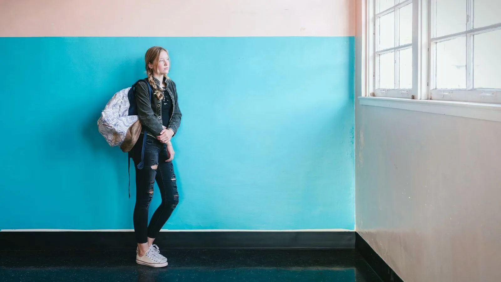 A teenage girl with a backpack standing alone against a bright teal wall