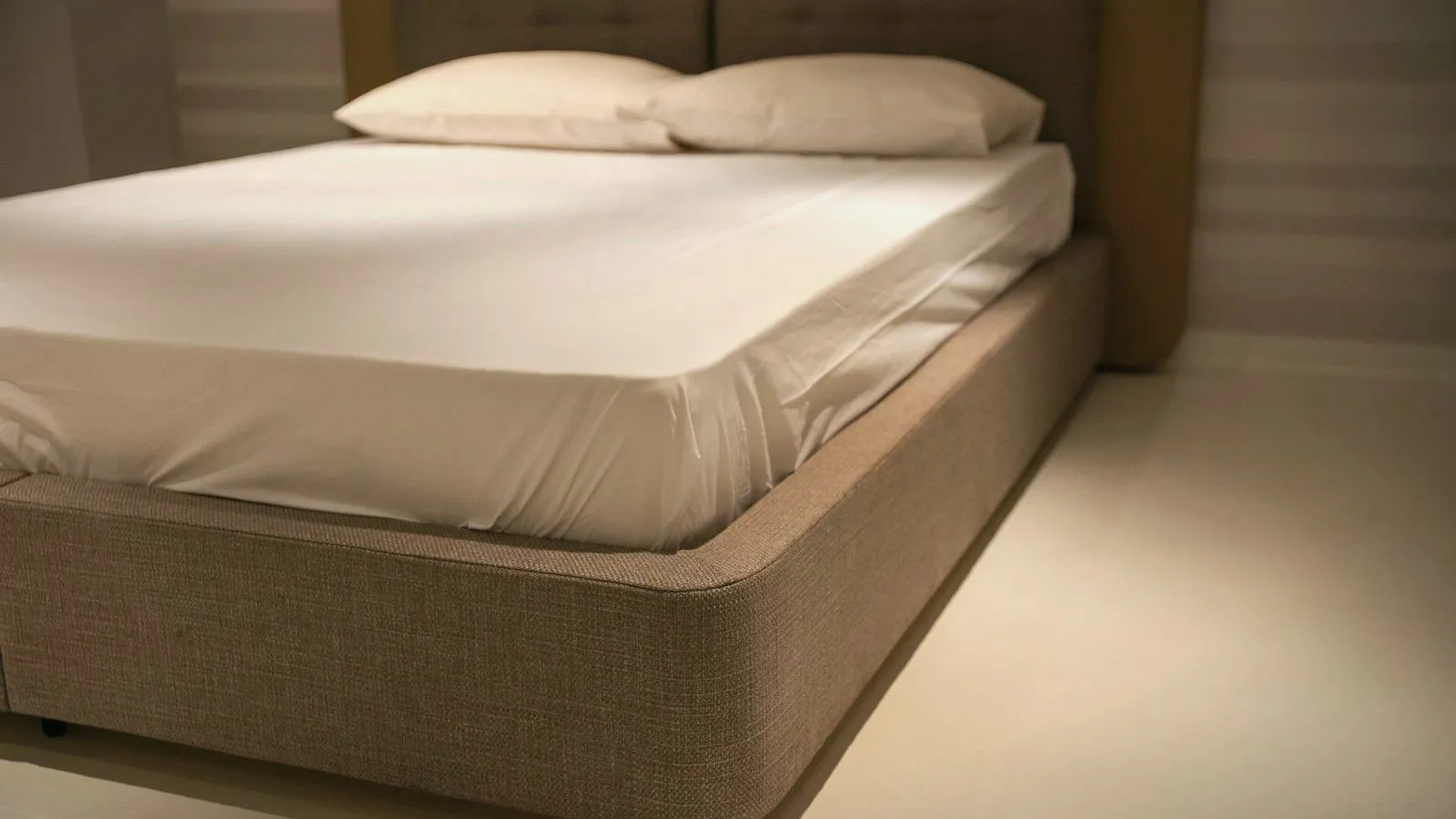 A bed with a white fitted sheet pulled tight over the mattress and two pillows, on a beige fabric base, seen from one corner.