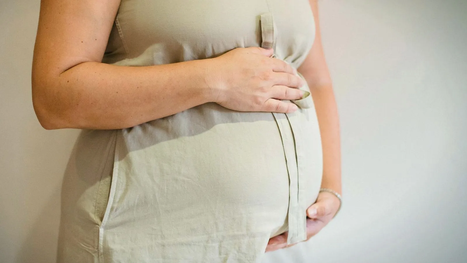 A pregnant woman in a pale linen dress stands by a white wall with one hand on top of the belly and one beneath it.