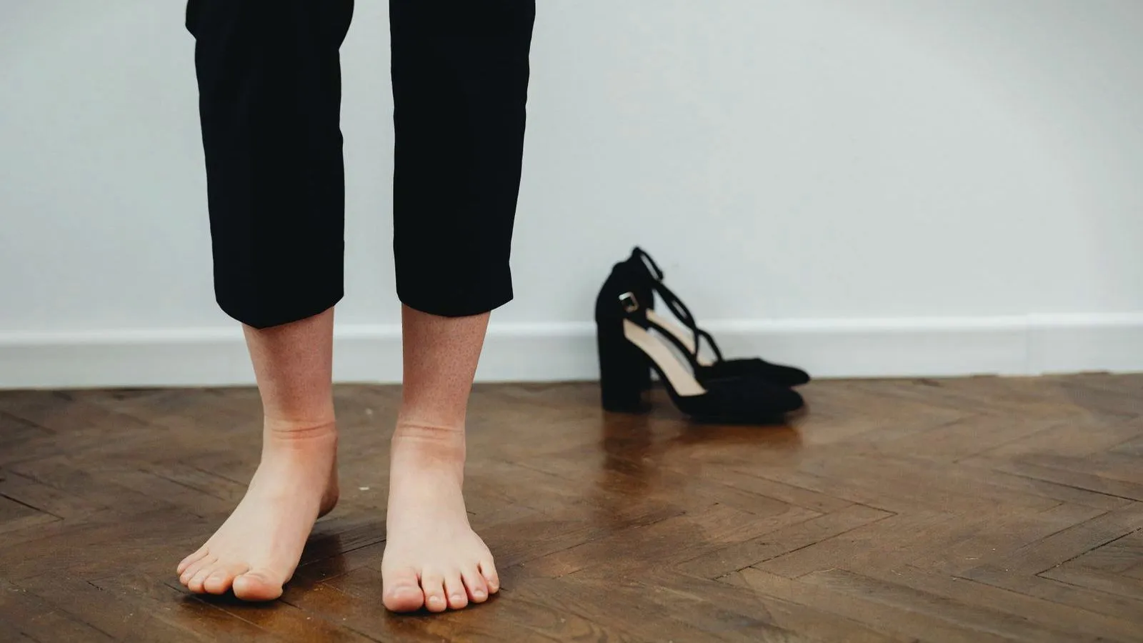 Bare feet standing on a wooden floor beside a discarded black high-heeled shoe