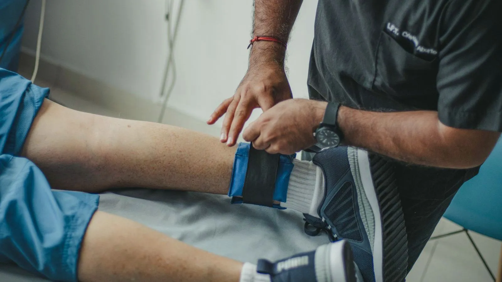 A physiotherapist in a dark uniform fastening a blue and black cuff around a patient's lower leg on a treatment couch