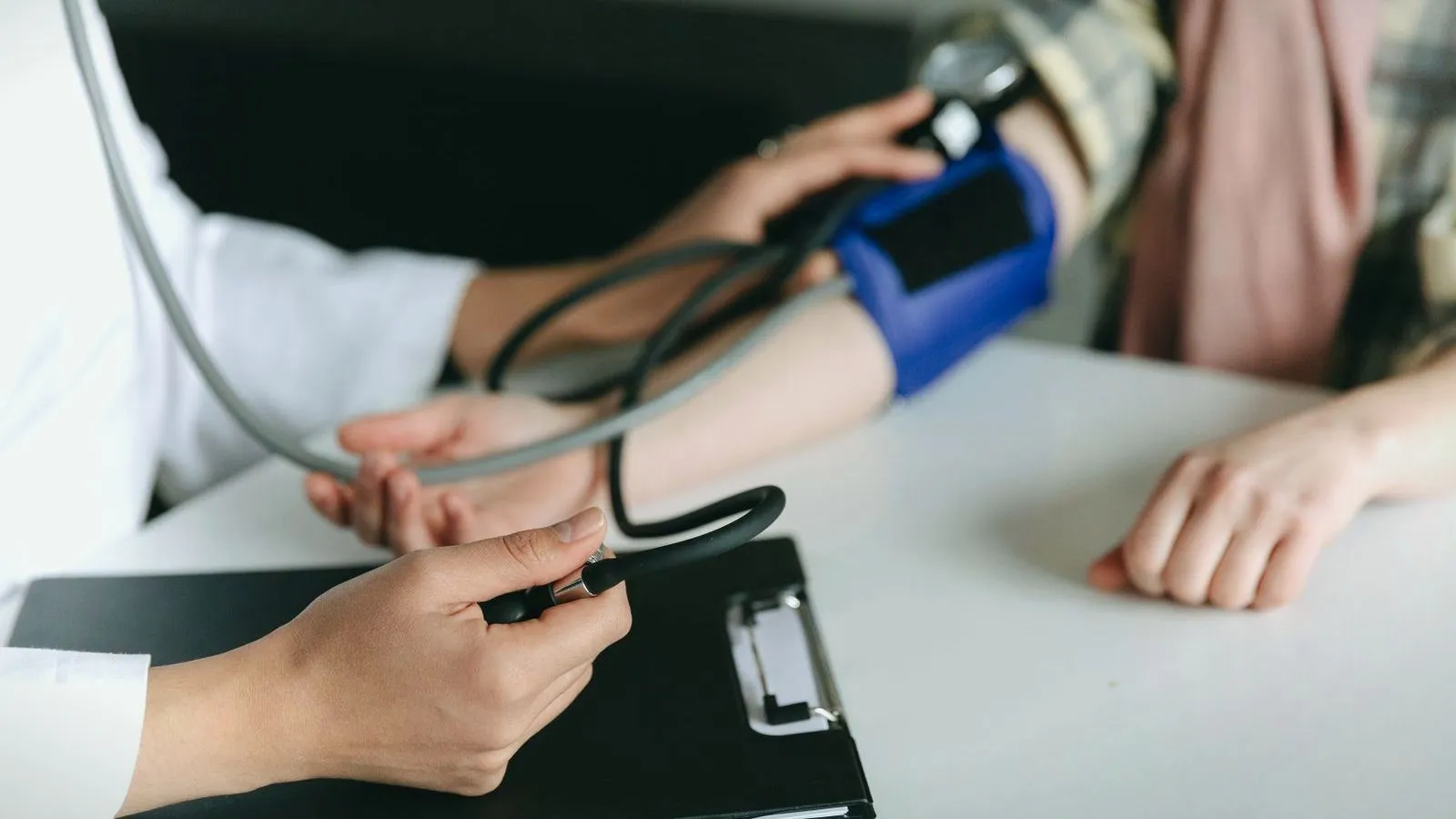 A clinician squeezes the bulb of a manual blood pressure cuff wrapped around a patient's arm resting on a white desk.