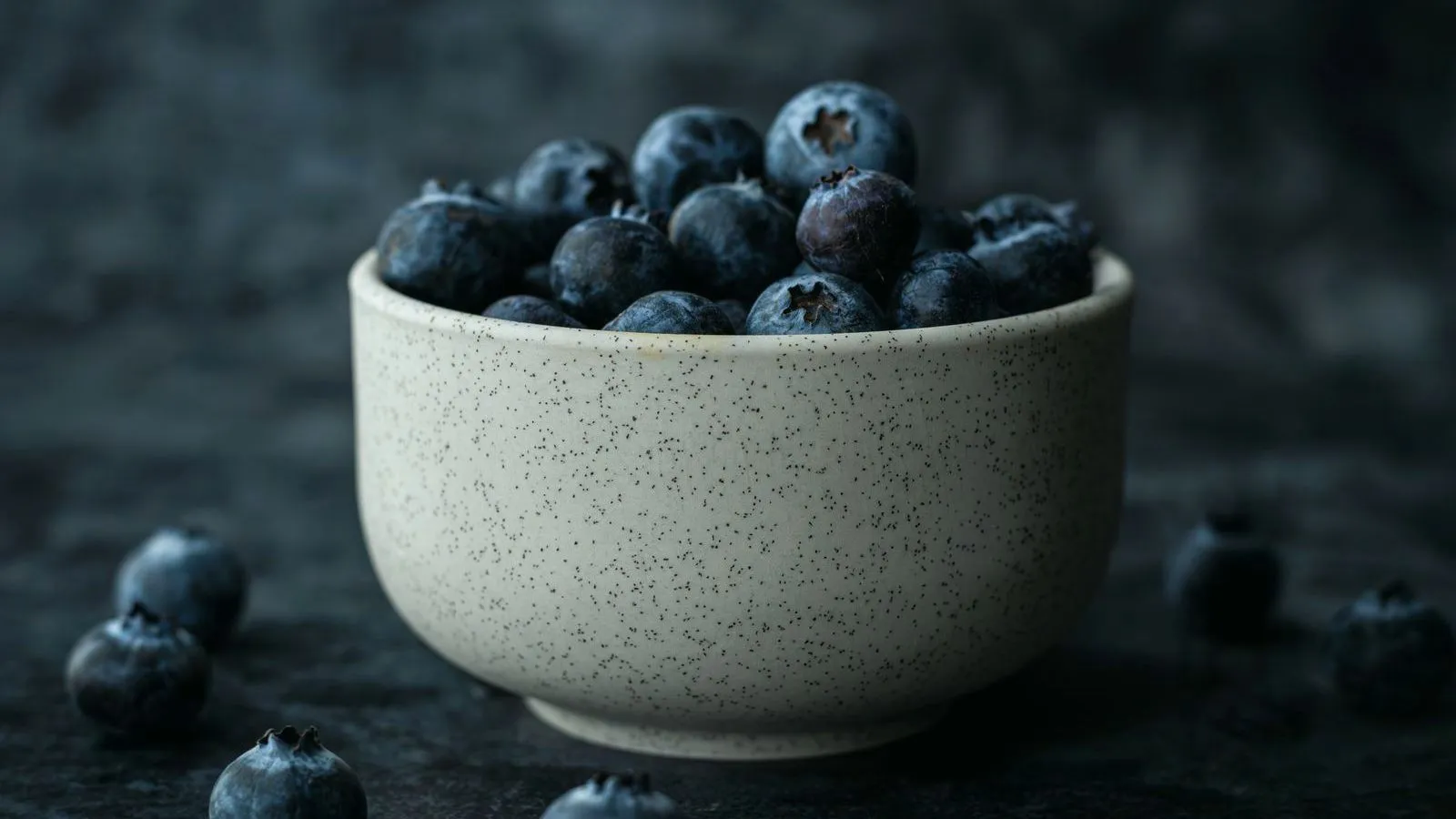 A speckled ceramic bowl heaped with blueberries on a dark surface, with a few loose berries around it.