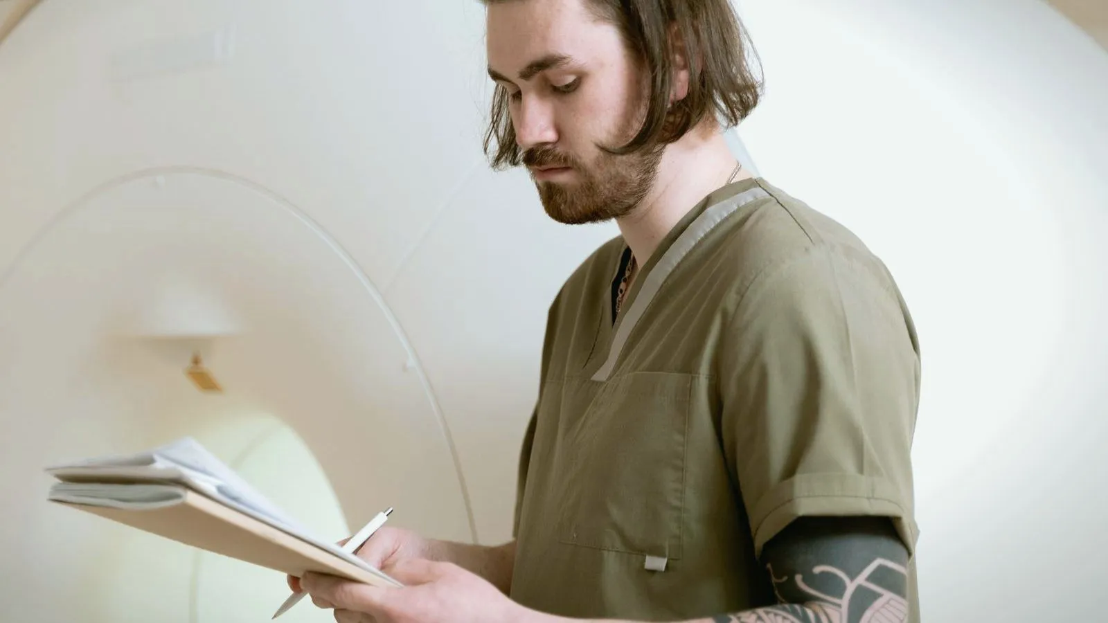 A person in olive scrubs writes on a sheaf of papers while standing in front of a large white scanner.