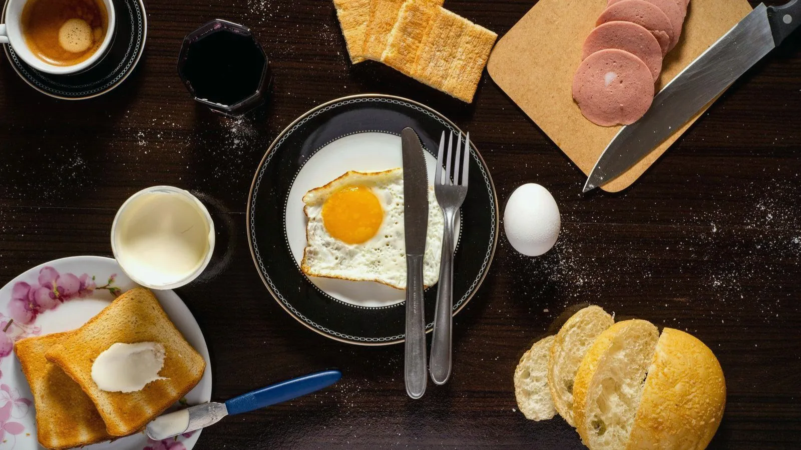 A breakfast table seen from above, with a fried egg, buttered toast, bread rolls, sliced sausage and coffee.