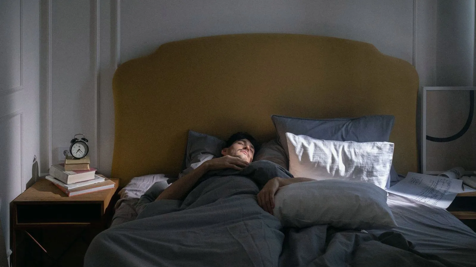 A man asleep on his back in a dim bedroom beside a bedside clock