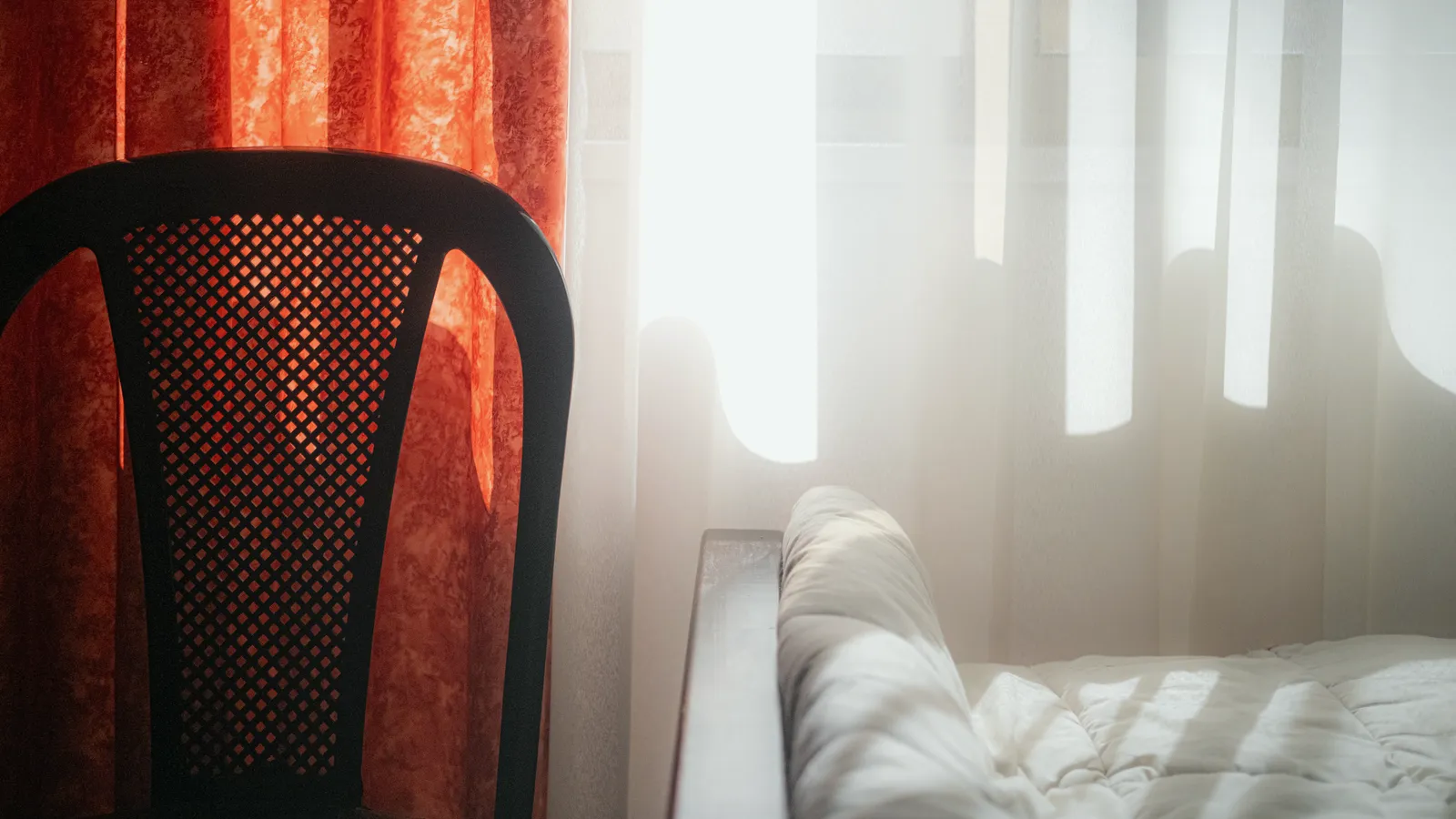 Bright daylight streaming through sheer curtains onto the corner of a bed beside a dark chair