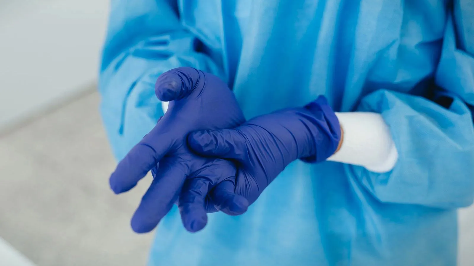 A person in a blue protective gown pulls on purple-blue medical gloves, seen from the chest down.