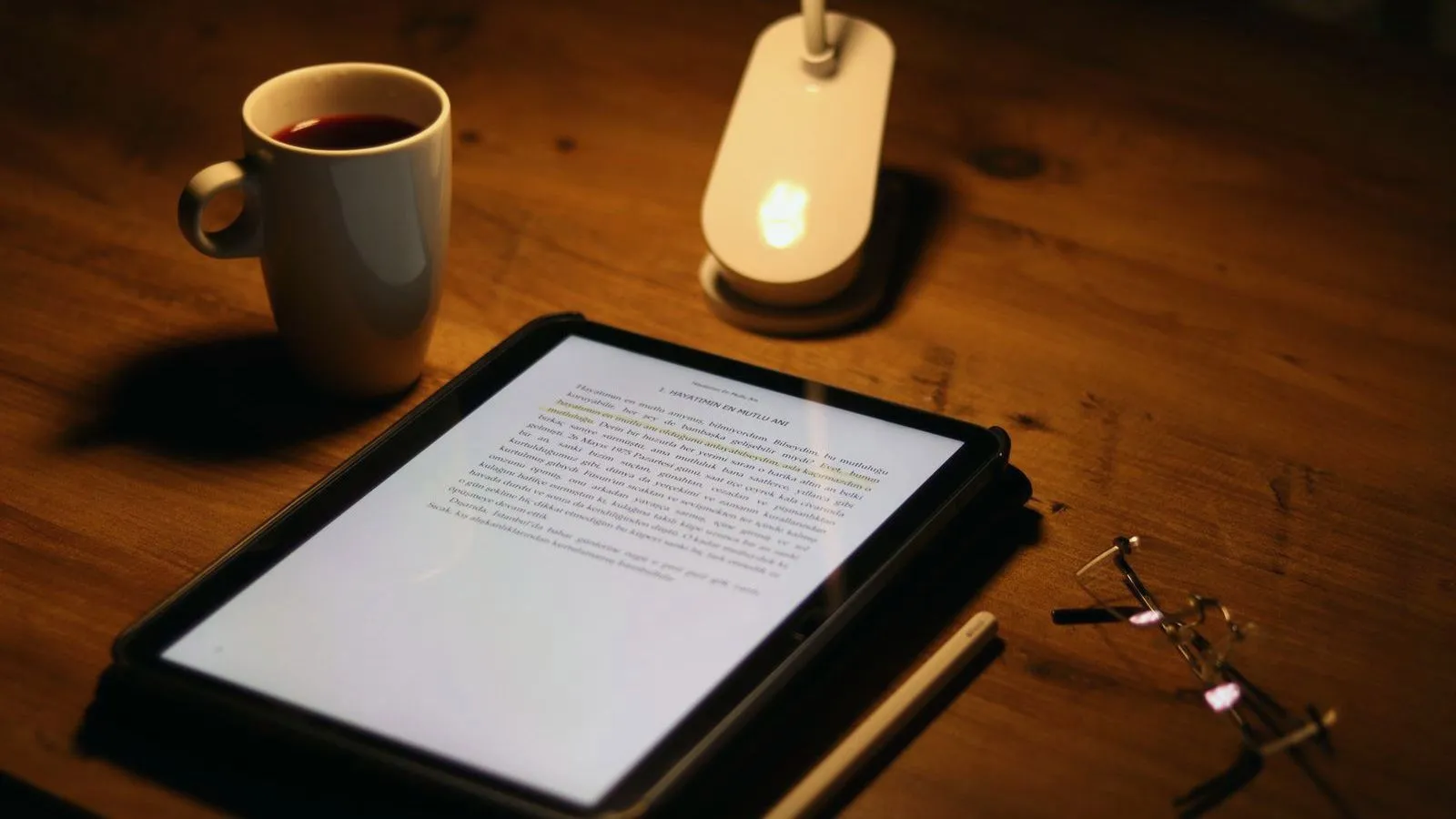 A mug of dark drink beside a lit tablet, a small lamp and a pair of glasses on a wooden table at night.