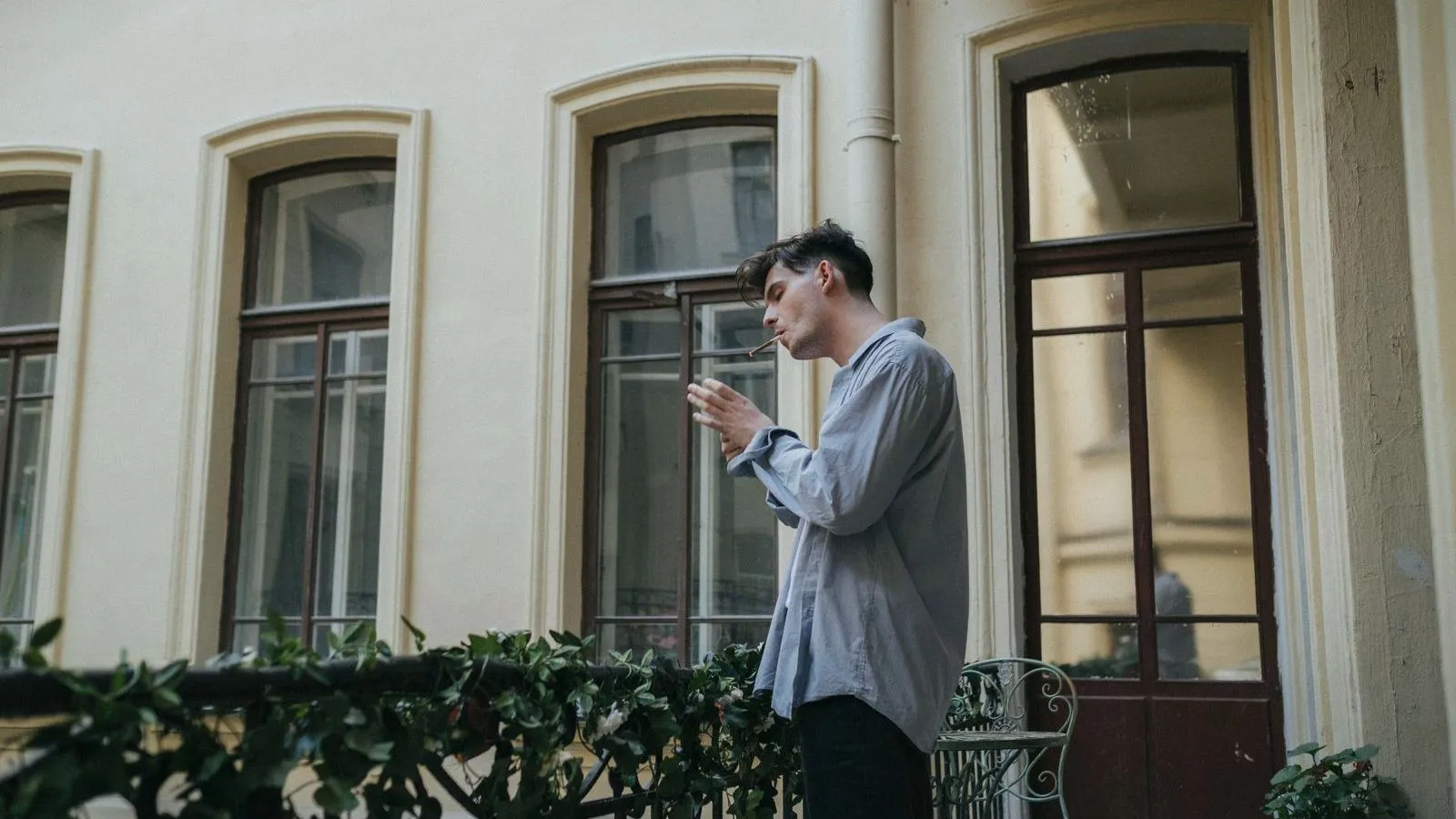 A person in a gray shirt smokes a cigarette on a balcony outside a pale building with tall arched windows.