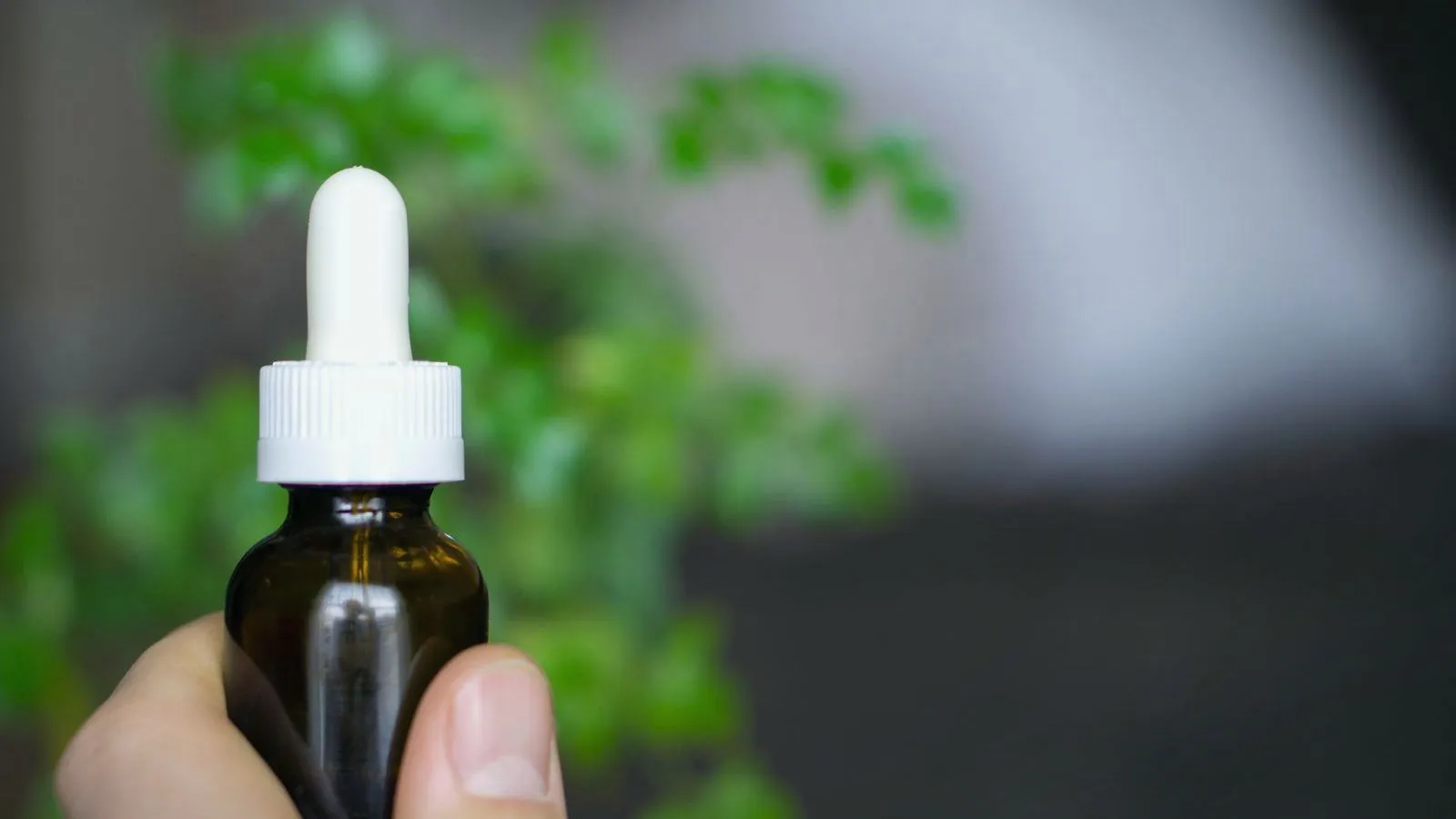 A hand holding an unlabelled amber dropper bottle with a white pipette, green leaves blurred behind