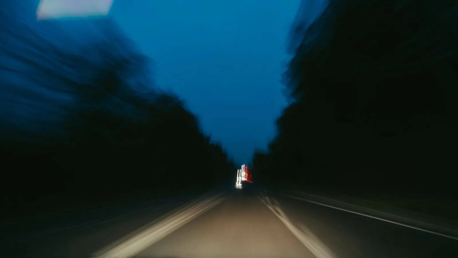A dark, motion-blurred road at dusk seen from a moving car, lined with trees