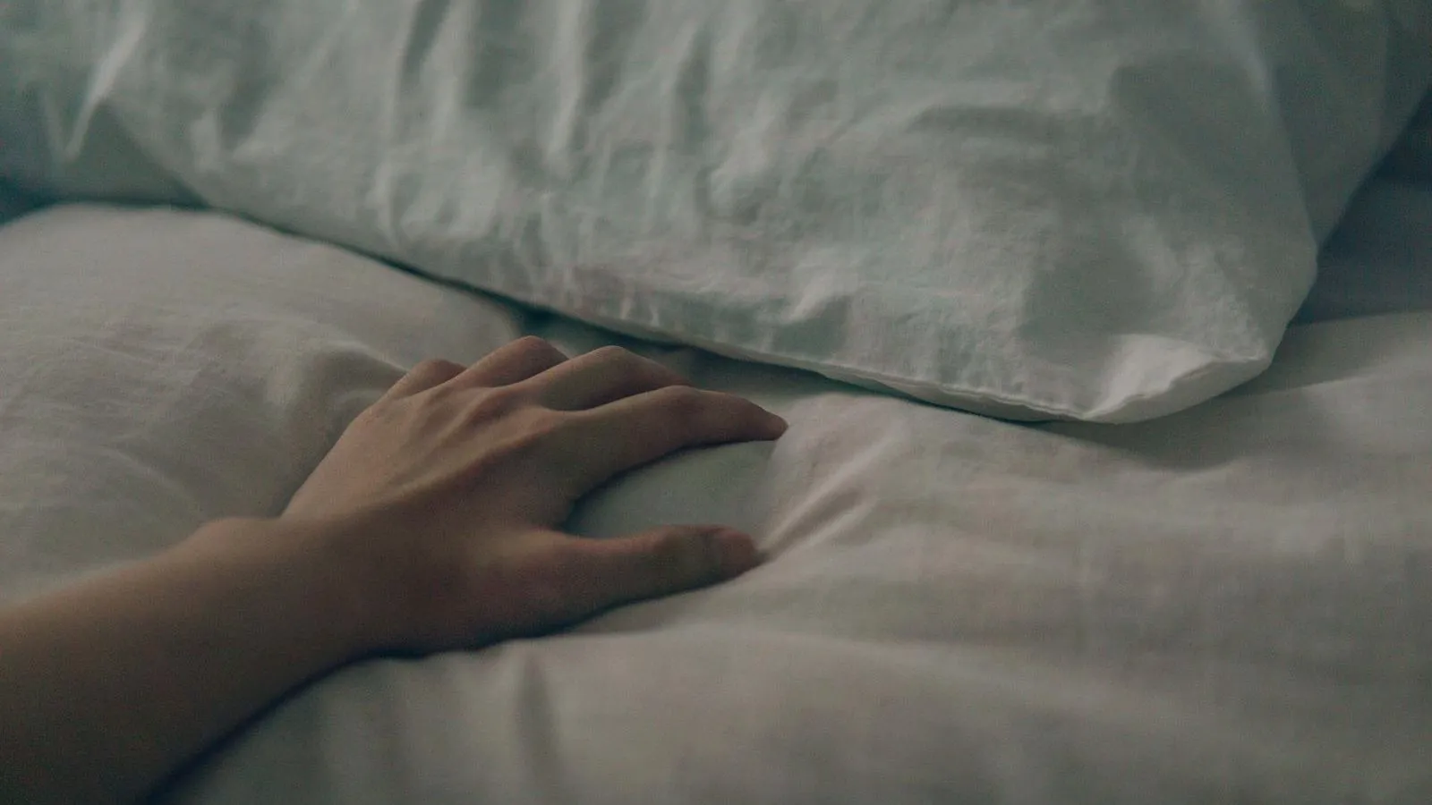 A hand rests on rumpled pale bedding beside a pillow.