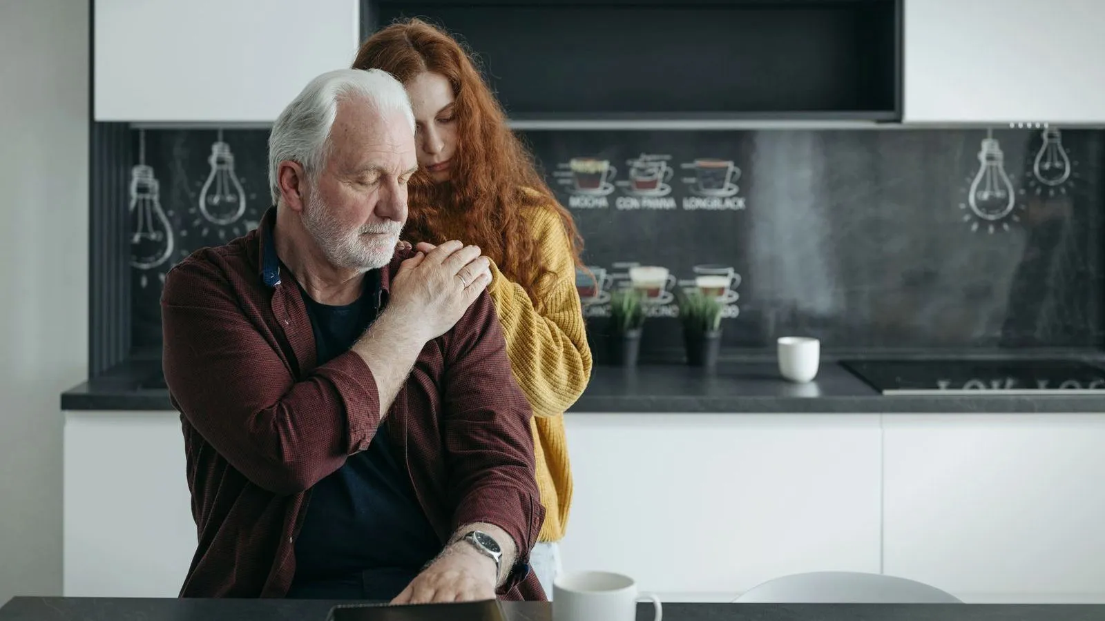 A young woman stands behind a seated older man with her arms around his shoulders, his hand resting on hers, in a kitchen.