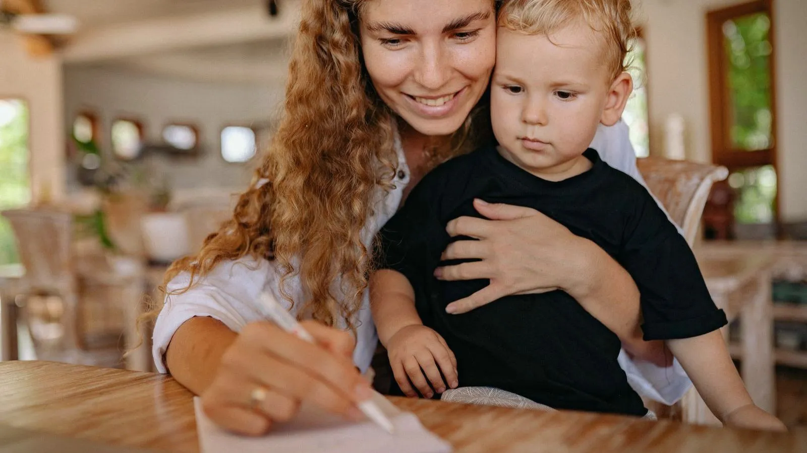 A smiling mother holding a toddler on her lap while writing at a table.