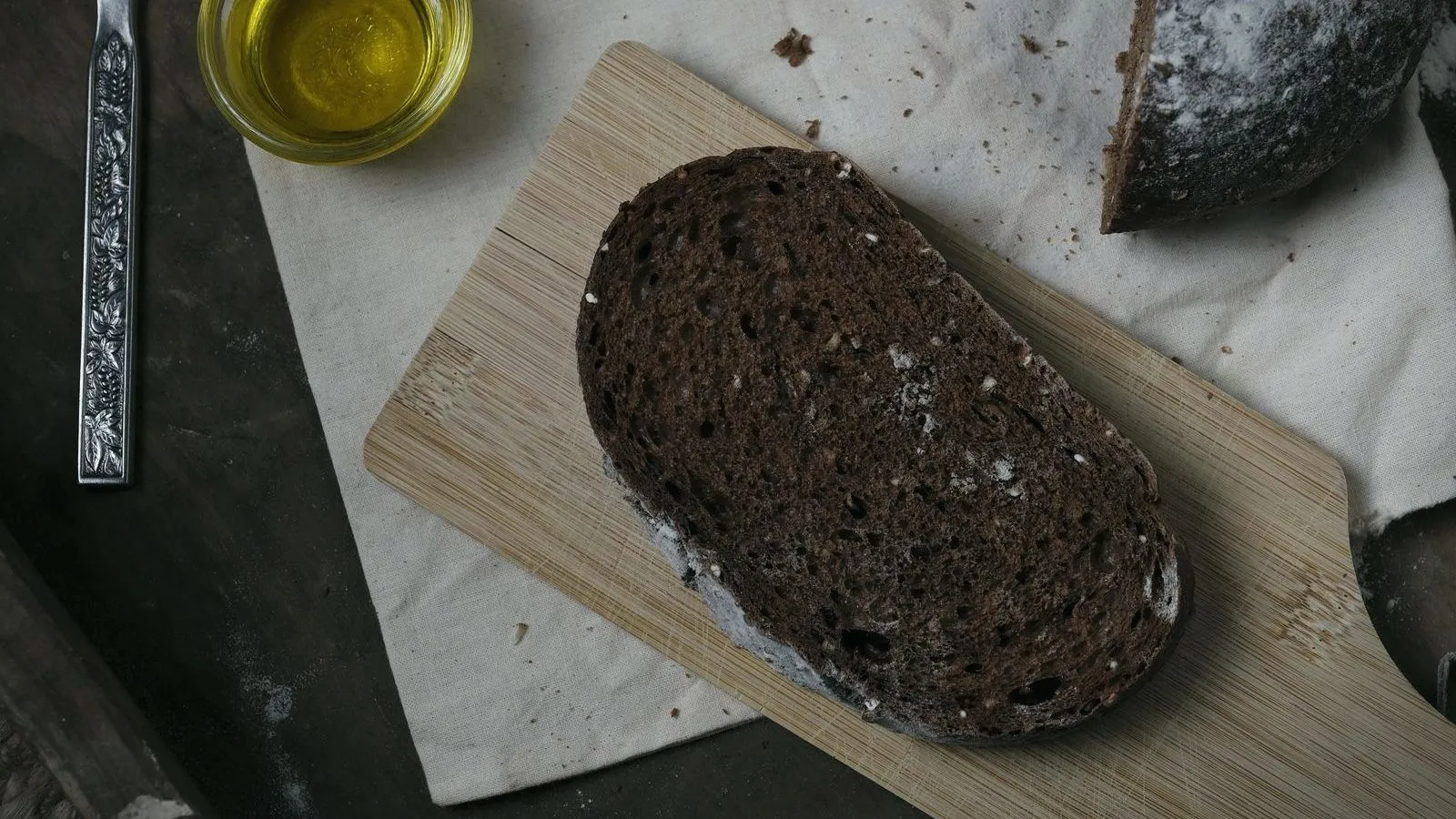 A dark rye loaf sliced on a wooden board with a small dish of olive oil