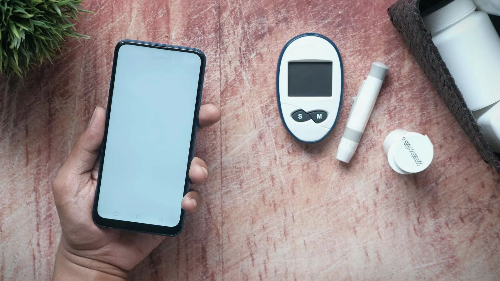 A hand holds a smartphone with a blank screen beside a glucose meter, a lancing device and a strip container on a wooden table.