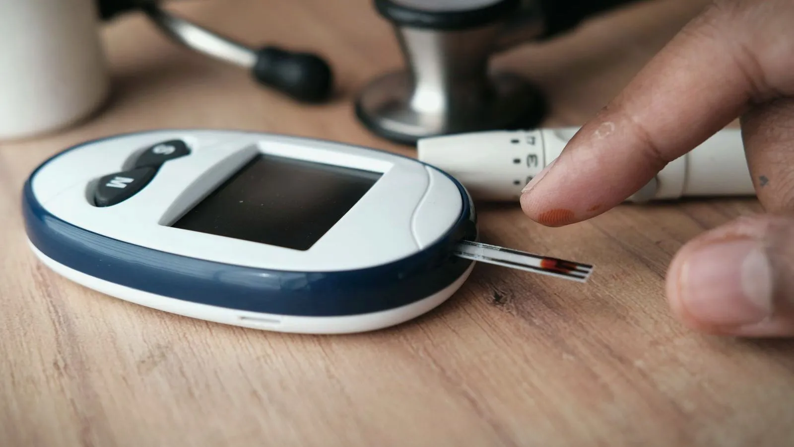 A blood glucose meter on a wooden table with a test strip holding a drop of blood, a fingertip and a lancing pen beside it.