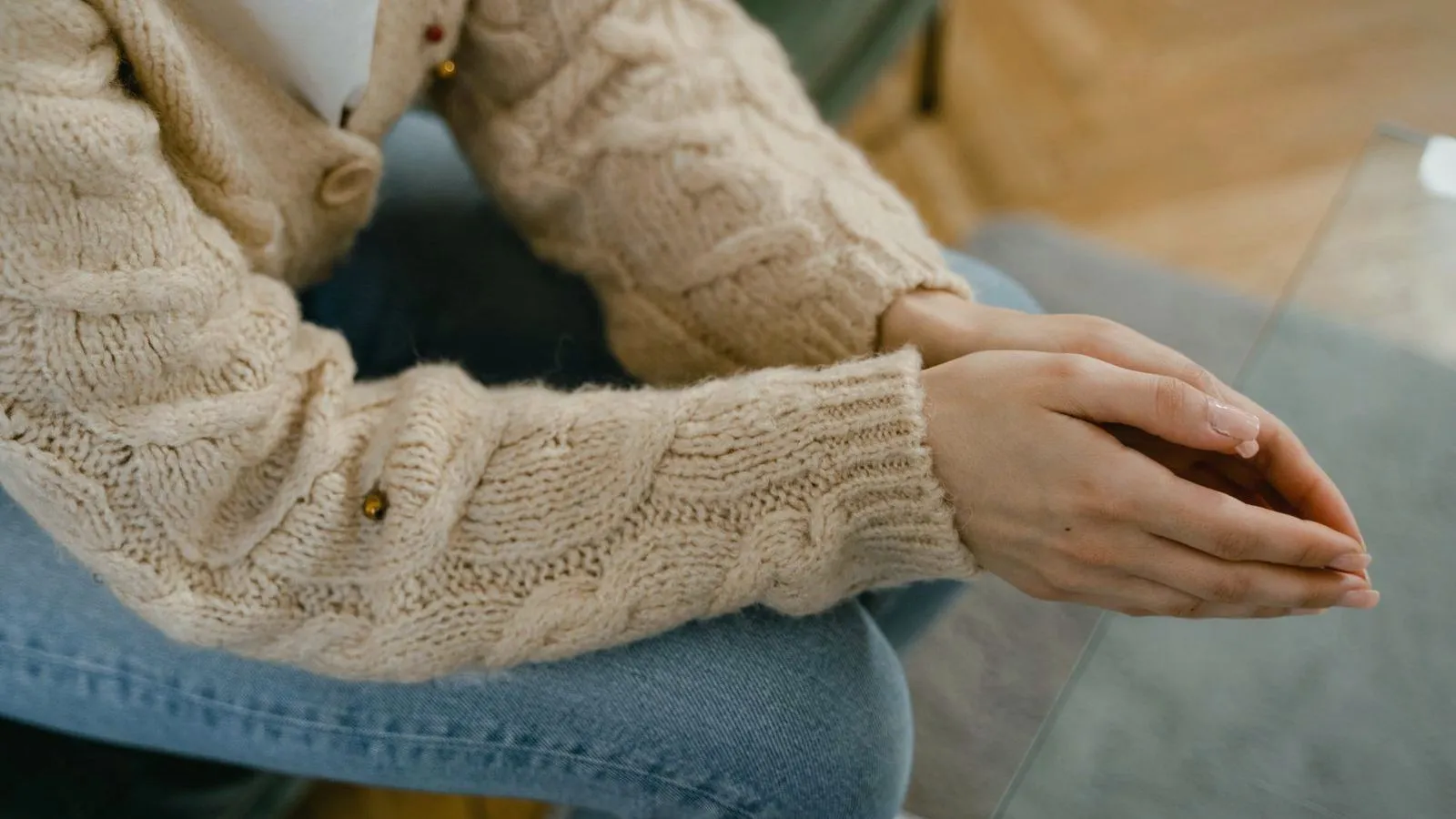 A person in a cream cable-knit sweater and jeans sits leaning forward with hands pressed together over the knees.