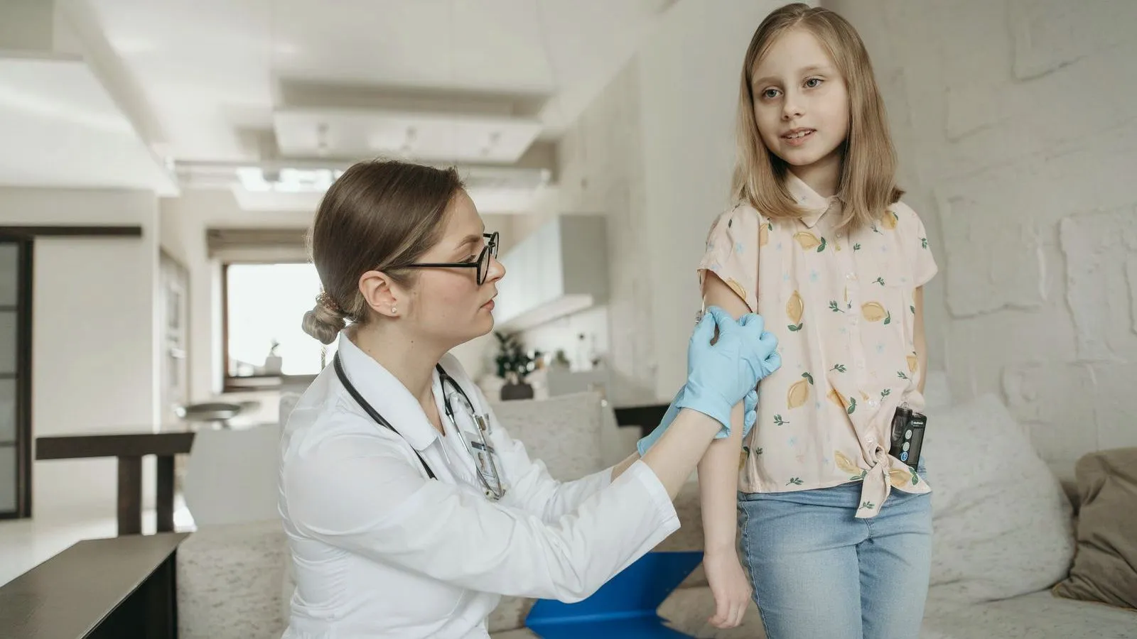 A clinician in a white coat and blue gloves holds the upper arm of a girl standing in a bright room.
