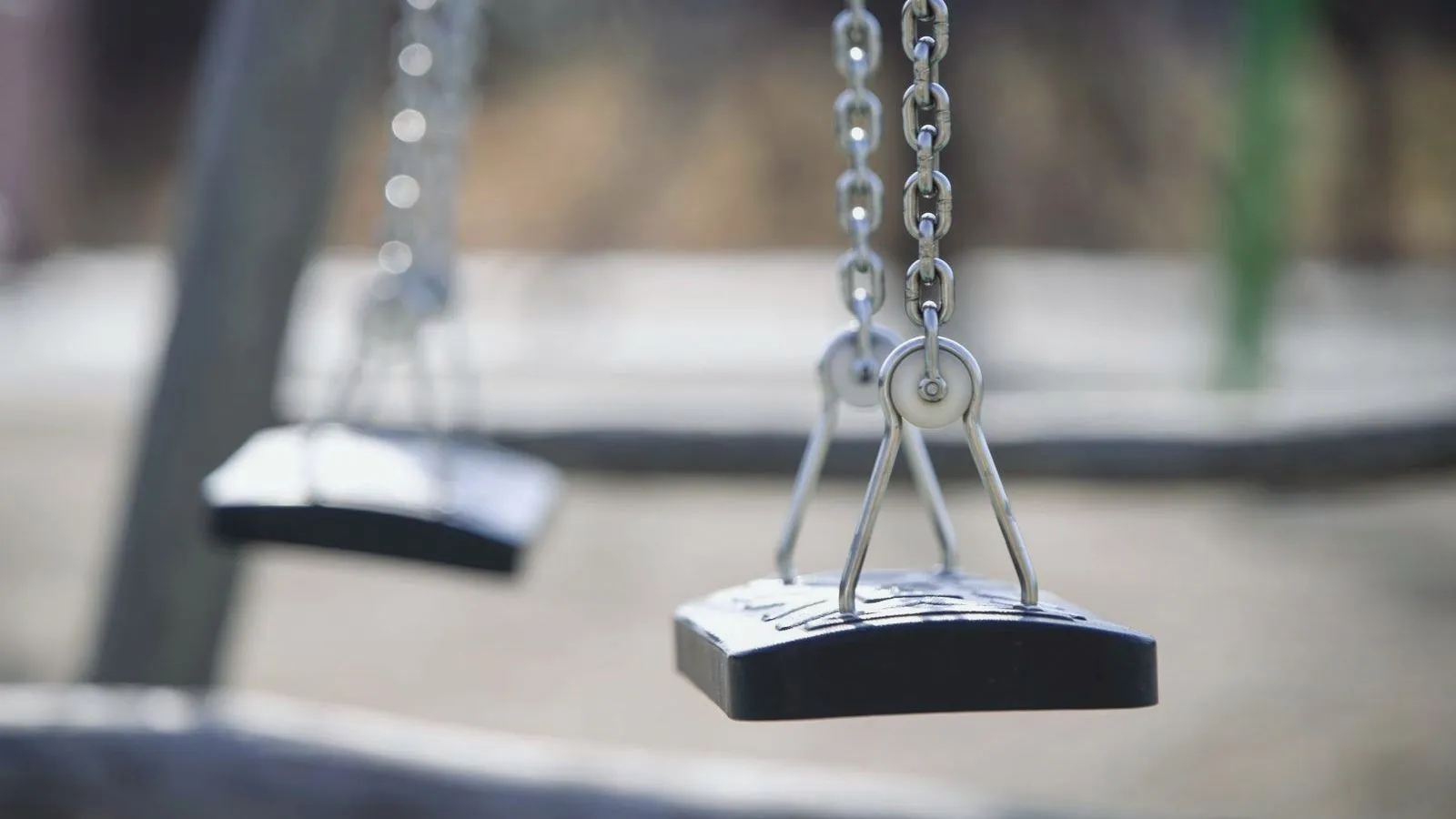 Two empty black swing seats hang from metal chains, with a blurred playground in the background.