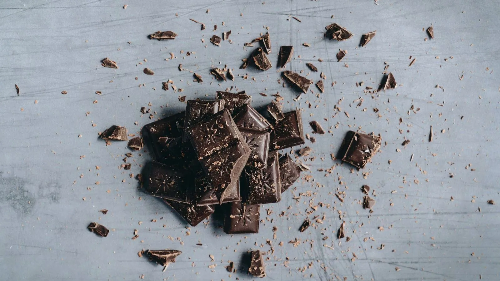 Broken pieces of dark chocolate scattered with cocoa crumbs on a grey surface