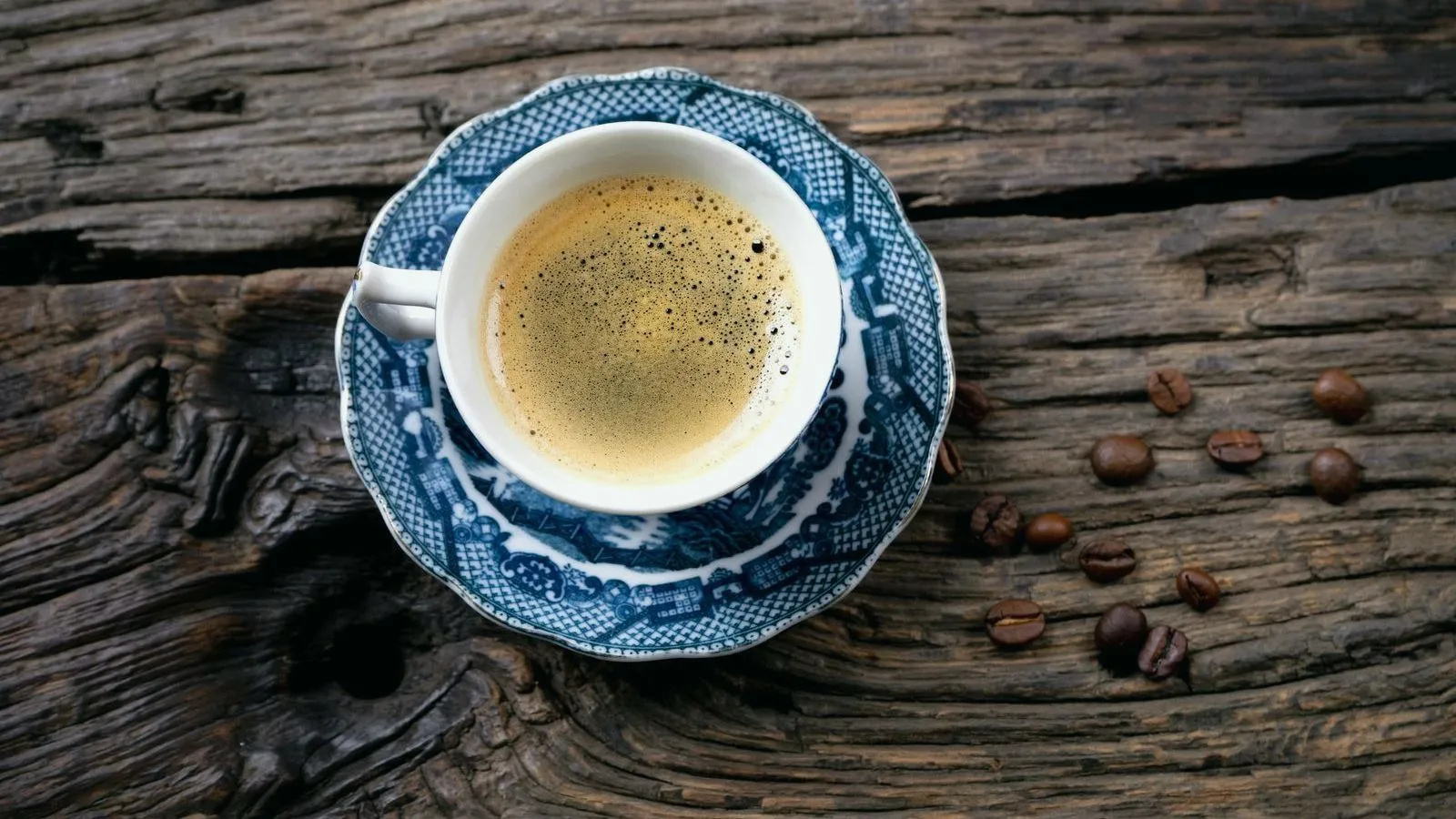 An espresso in a blue and white cup on weathered wood beside coffee beans