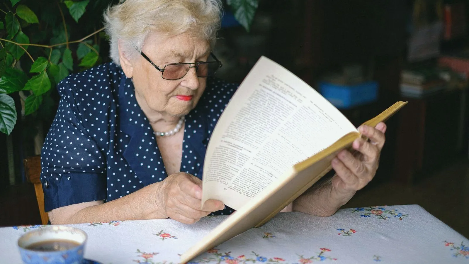 An older woman in glasses reading a book at a table beside a cup and saucer