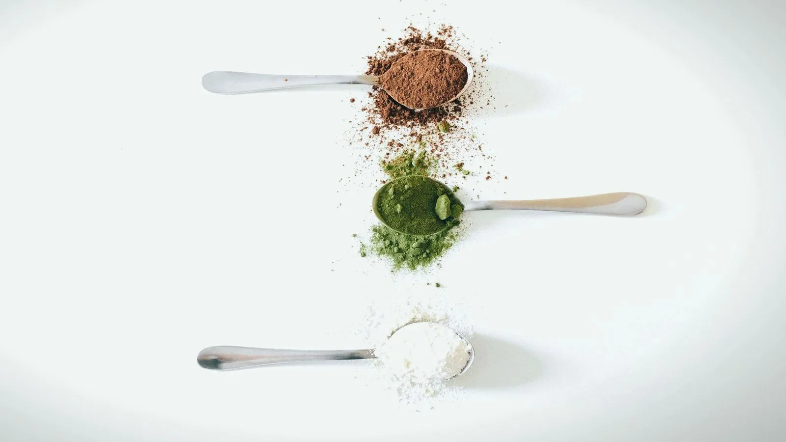Three spoons of powder, brown, green and white, on a white surface.