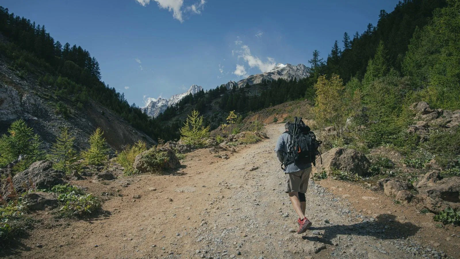 A backpacker seen from behind walks up a gravel track through a mountain valley with snowy peaks under blue sky.