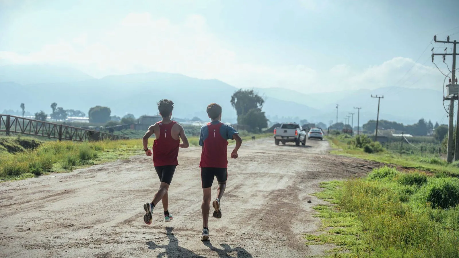 Two teenagers in running vests run away from the camera down a dirt country road.