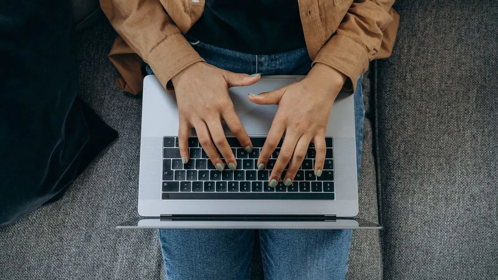 Two hands type on a laptop resting on the lap of a person in jeans and a tan jacket, seen from above.