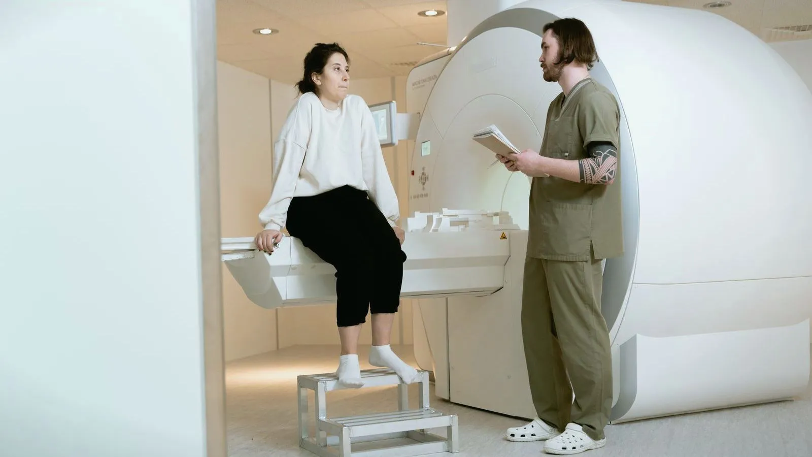 A patient sitting on the table of a large medical scanner, talking with a technician who holds a tablet.